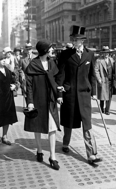 Man and woman walking on a New York City street in the late 1920s, both holding lit cigarettes, dressed in formal period clothing